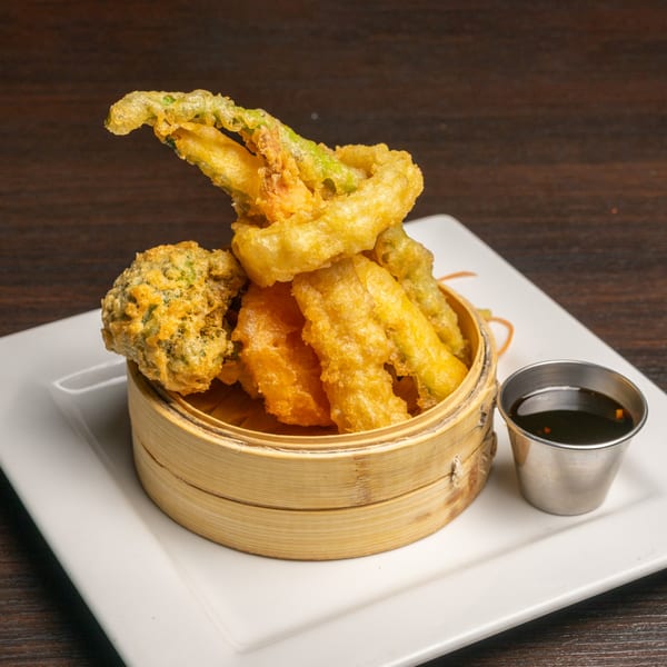 Crispy vegetable tempura, including broccoli, in a bamboo steamer with a side of dipping sauce.