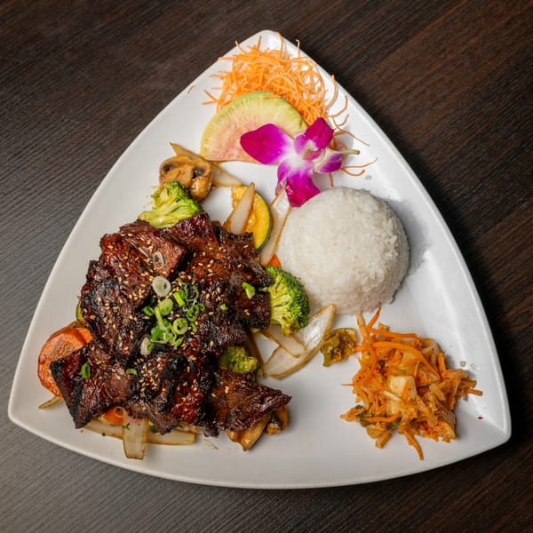 Grilled short ribs with rice, vegetables, and kimchi, garnished with an orchid on a white plate.