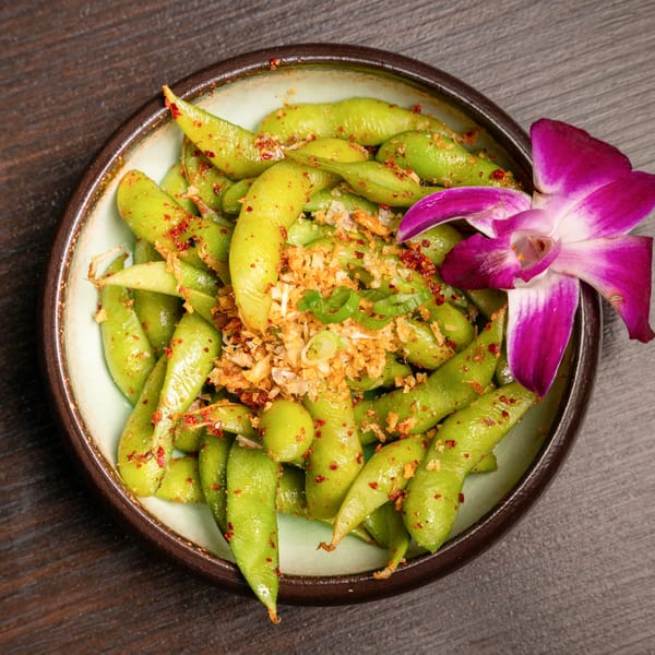 A bowl of spicy edamame with garlic and green onions, garnished with a purple orchid.