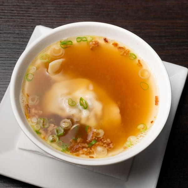 A white bowl of Japanese wonton soup with scallions and fried garlic on a dark table.