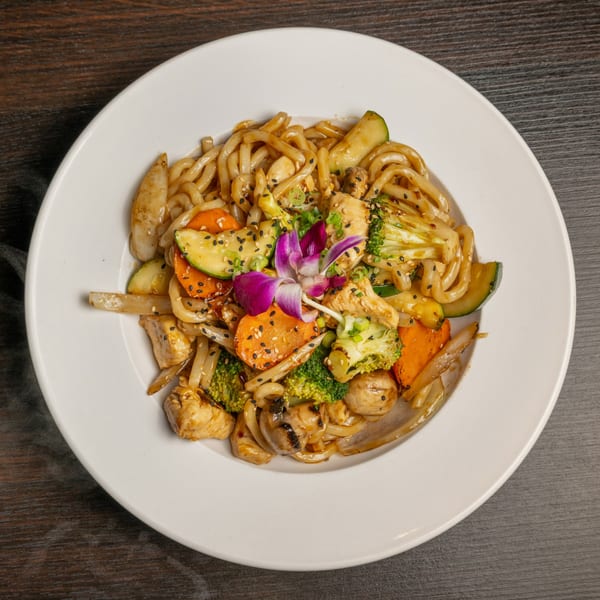 Udon noodles with chicken, broccoli, carrots, zucchini, mushrooms, and a purple orchid garnish.